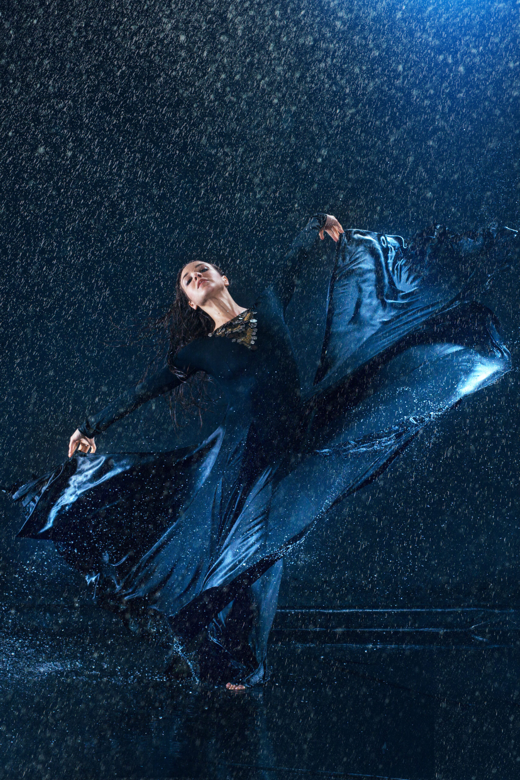 The young beautiful modern dancer in long dress dancing under water drops in blue aqua studio